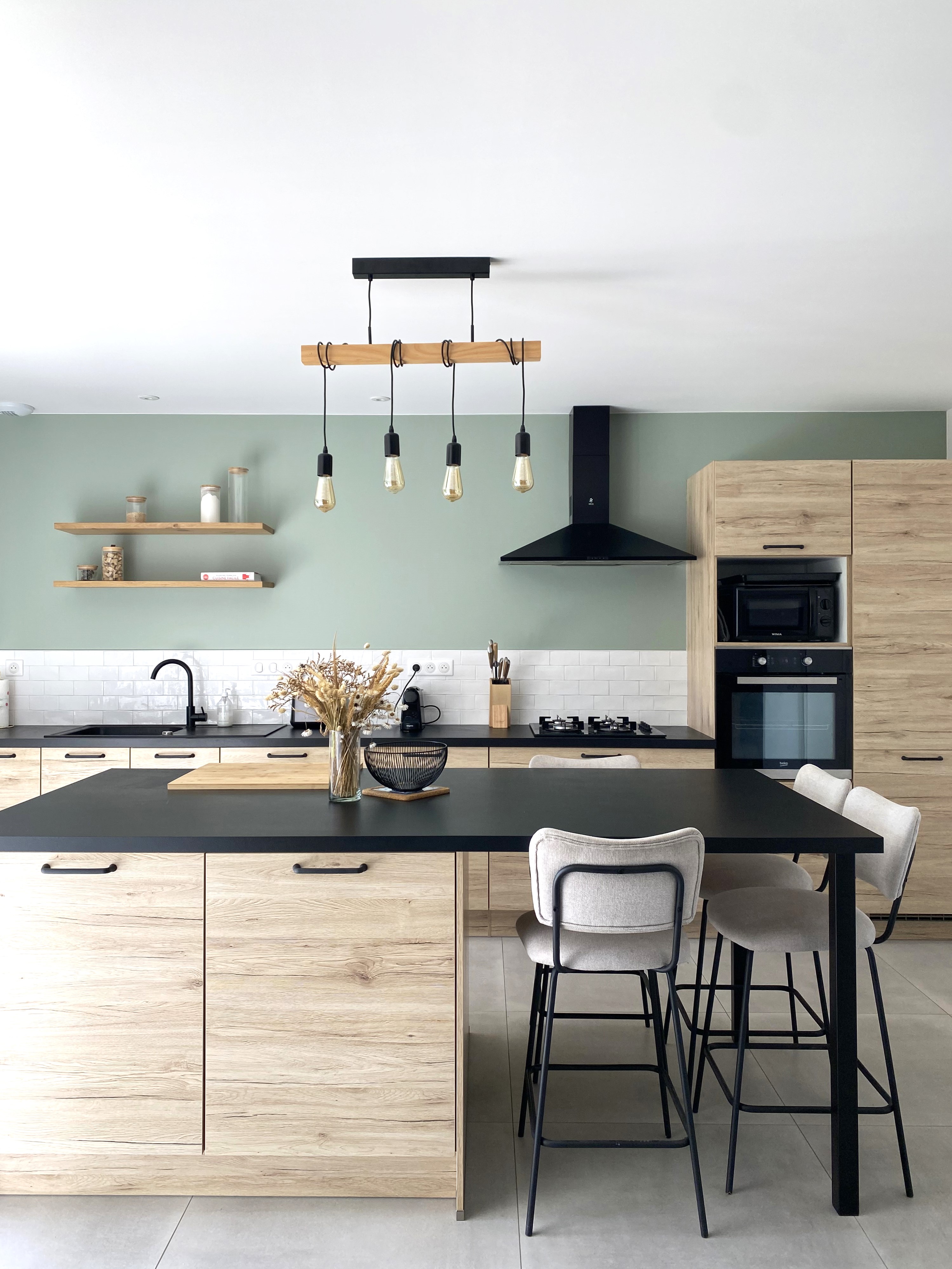 Cuisine rénovée mur vert sauge avec façades bois, plan de travail stratifié noir, ilôt central et chaises bar tissu gris — Projet Tremblay, architecture d’intérieur à Rennes